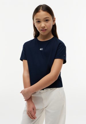 Κορίτσι με μακριά σκούρα μαλλιά φοράει εφαρμοστή μπλούζα σε ναυτικό μπλε με κοντά μανίκια και άσπρο παντελόνι, στέκεται με τα χέρια χαλαρά σταυρωμένα μπροστά.