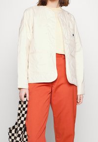 Prošívaná krémová bunda s kulatým límcem, zapínáním na patentky a přední kapsou, v kombinaci s oranžovými kalhotami a taškou s černobílým vzorem.