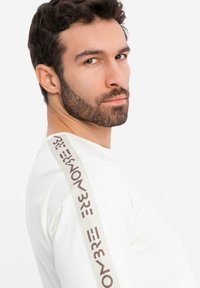 Muž s krátkými kudrnatými vlasy a vousy, oblečený v bílé košili s n ápisem "3NOM3" na pásce na rameni, dívající se přes rameno na bílém pozadí.