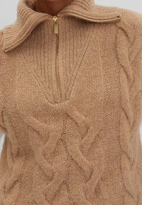 Pull en maille marron avec de larges motifs torsadés et une fermeture éclair dorée partiellement ouverte au niveau du col haut et plissé.