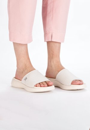 Sleek white slide sandals with a soft-textured upper and an open toe design, featuring a subtle logo and cushioned sole, paired with pink cropped pants.