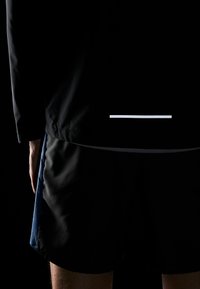 Zwarte lichtgewicht jas heeft een reflecterende witte horizontale streep op de achterkant. De shorts zijn zwart met een gladde textuur en blauwe zijaccenten.