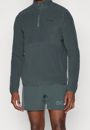Mężczyzna ubrany w ciemnozieloną polarową bluzę z zamkiem do połowy długości i dopasowane szorty, oba z logo "BORG" na piersi i nodze.