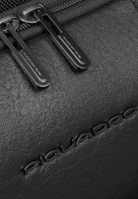 Material de cuero texturizado negro que muestra una cremallera con dos tiradores metálicos. Presenta el logo en relieve "Piquadro", resaltando un diseño elegante.