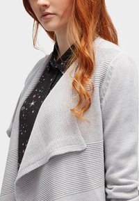 Jasnoszara kardiganka z szalem o teksturowanym wzorze dzianiny. Zaprojektowana z opadającym przodem. Noszona na czarnej koszulce z wielokolorowymi wzorami przypominającymi gwiazdy.