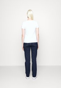 Woman with long blonde hair wearing a white short-sleeve shirt, dark blue jeans, and white sneakers standing facing away on a plain background.