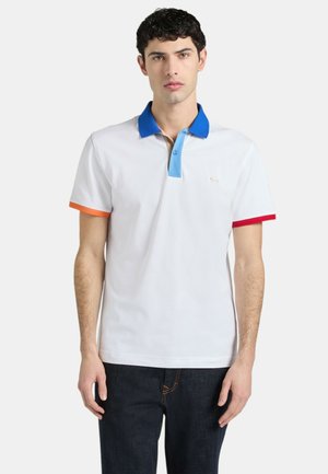 Giovane uomo che indossa una polo bianca con colletto blu, pattina azzurra, bordo manica sinistro arancione e bordo manica destro rosso, abbinata a jeans scuri.