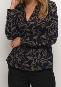 Blouse marine à manches longues avec motif floral beige, encolure en V et poignets boutonnés ; texture lisse, coupe ajustée, longueur aux hanches.