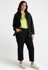 Czarna zip-up kurtka noszona na jasnej zielonej bluzie, w zestawie z czarnymi spodniami i białymi sneakersami. Posiada luźny krój i prosty design.