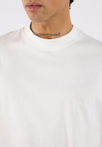 White cotton t-shirt with a crew neckline, smooth texture, and short sleeves. Displays a tattoo reading "believe in yourself" on the neck.