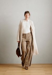 Lichtbeige gebreide cardigan over een witte blouse, gecombineerd met bruin geruite wijde broek en bruine enkellaarsjes, met een bruine tas.