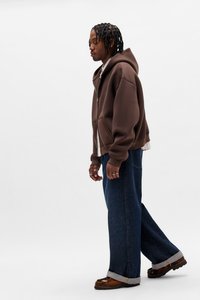 Felpa marrone con zip sopra una maglietta bianca, abbinata a jeans denim scuri a gamba larga e scarpe marroni. L'outfit presenta un tessuto strutturato e orli risvoltati.