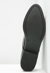 Semelle de chaussure noire avec un fond lisse et plat, présentant une texture à motifs et un design à découpe ronde. Étiquette de taille visible près du talon.