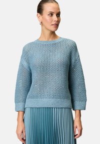 Maglione azzurro chiaro a maglia, con una trama ampia e aperta, caratterizzato da un collo rotondo e maniche a 3/4, abbinato a una gonna blu plissettata.