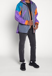 Grå vindjacka med färgglada accenter i teal, orange och lila. Har en dragsko huva och dragkedjefickor. Bärs med mörka jeans och svarta skor.