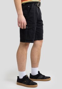 Sorte cargo shorts lavet af bomuld med flere lommer, knapdetaljer og en afslappet pasform. Bæres med sorte sneakers og hvide ankelstrømper.