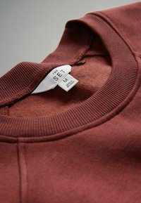Bordeauxrode sweater met een geribbelde ronde hals en een zachte textuur. Voorzien van een wit label met "THE SET" en maat "M" aan de binnenkant van de hals.