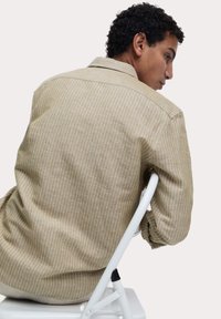 Tan gestreept shirt met een zachte textuur, een knoopsluiting bij de kraag en een relaxte fit, gezien vanuit de achterkant terwijl het model op een witte stoel zit.