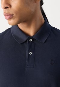 Polo shirt bleu marine en tissu texturé, avec une patte de boutonnage à deux boutons et un logo discret sur la poitrine. Présente des boutons contrastés et un col classique.
