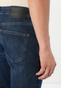 Jeans de mezclilla de color azul oscuro con un parche de cuero con texto en relieve en la cintura y un bolsillo trasero con detalles de costura estándar.