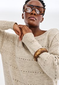 Pull en tricot beige avec des lignes texturées horizontales et de petits trous, associé à de grandes lunettes de soleil en écaille de tortue et un bracelet en bois.
