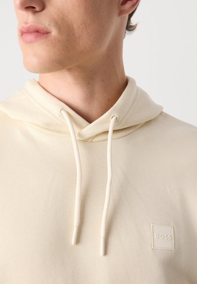 Hoodie crème en tissu texturé, présentant une capuche avec cordon de serrage et un patch logo carré avec "BOSS" sur la poitrine.