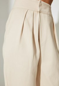 Pantalons plissés beige clair en tissu doux. Dotés d'une taille haute et d'une texture subtile. Coupe ajustée avec des lignes épurées.
