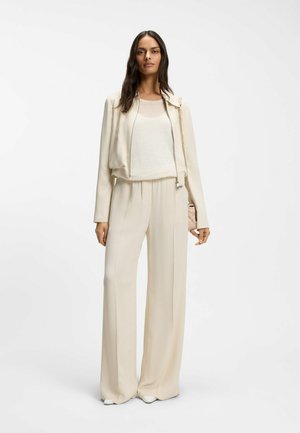 Pitkähiuksinen tummahiuksinen nainen, jolla on päällään kermaisen värinen takki, väljiä housuja ja vaalea toppi, kantaen beigen väristä ketjuhihnaista laukkua.