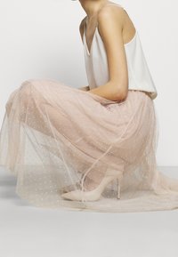 Jupe en tulle rose blush superposée à un jupon, ornée de pois blancs. Portée avec un débardeur crème et des chaussures à talons nude.