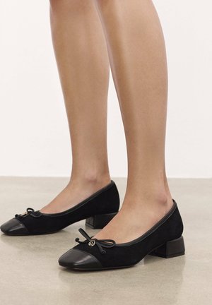 Black low-heeled shoes with a suede body, leather toe cap, and bow detail on the front. Includes a small gold accent on the bow.