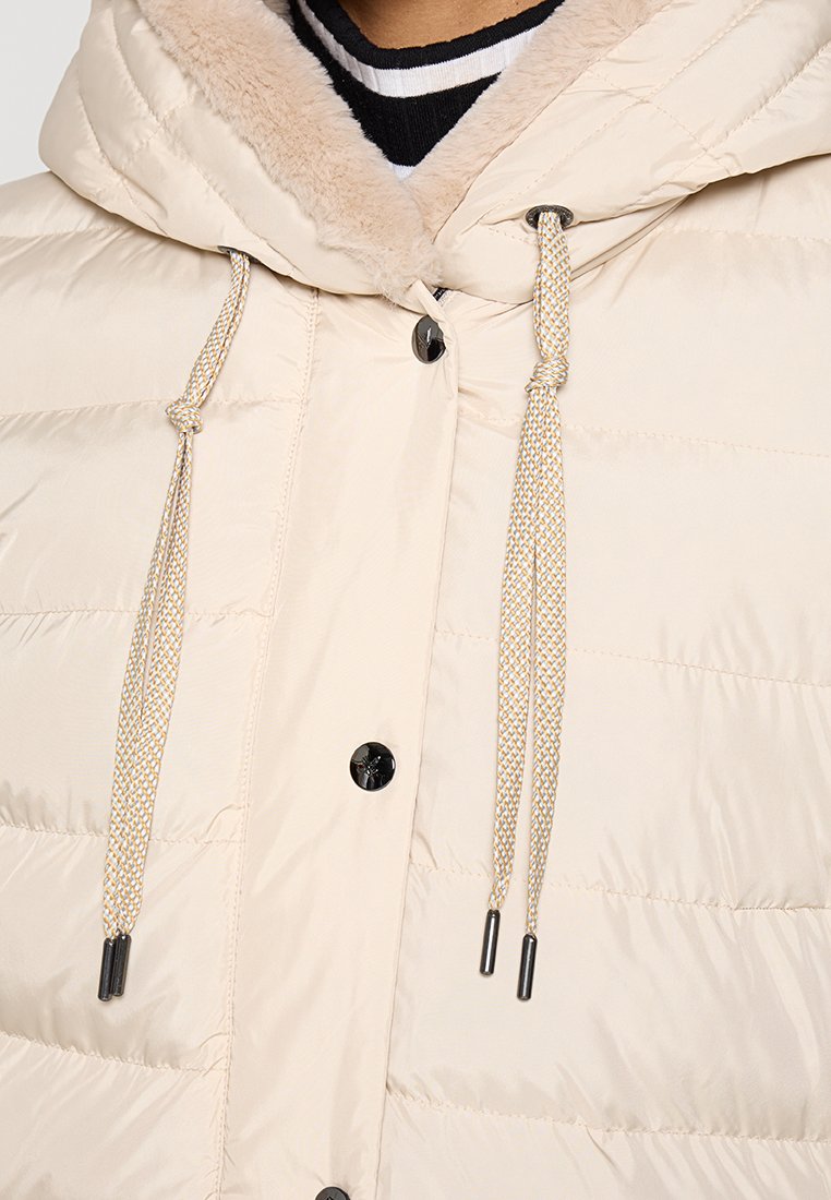 Kremowa kurtka puchowa z kapturem wyłożonym sztucznym futrem, z czarnymi napami i regulowanymi sznurkami z metalowymi końcówkami.