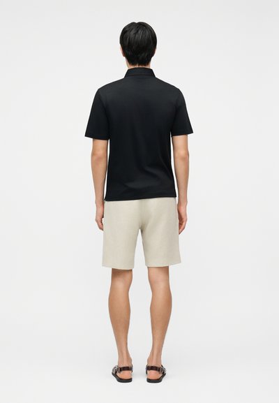 Άνδρας που φοράει μαύρη κοντομάνικη πουκαμίσα με γιακά, ανοιχτό μπεζ σορτς και μαύρα σανδάλια, στέκεται πλάτη στον θεατή μπροστά σε λευκό φόντο.