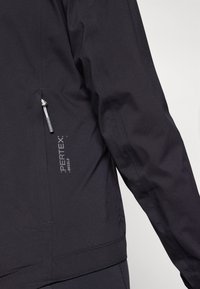 Svart Pertex-jakke med glidelåslomme, teksturert stoff og en tettsittende design. Inkluderer merkevaredetalj på ermet.