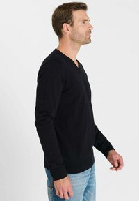 Zwarte V-hals trui met geribbelde schouderdetails, lange mouwen en een slanke pasvorm, gecombineerd met spijkerbroek. Soepele textuur, klassiek ontwerp.