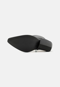 Chaussure à talon haut noire avec une pointe fine et une semelle en caoutchouc striée, présentant un dessus en cuir lisse. La semelle a une légère courbe pour plus de stabilité.
