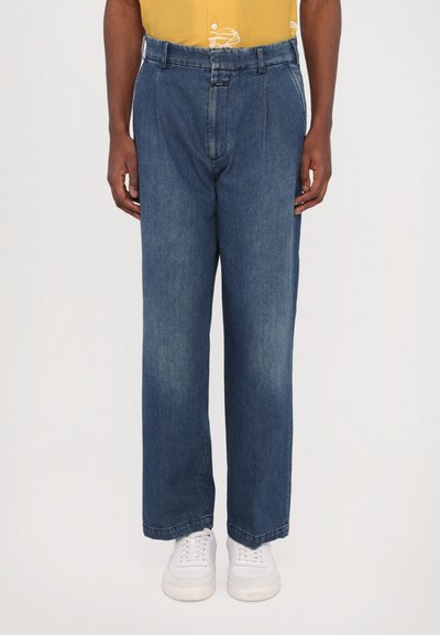 Blue denim trousers with a relaxed fit, featuring pleats, a button closure, and a contrasting waistband. Worn with white sneakers.