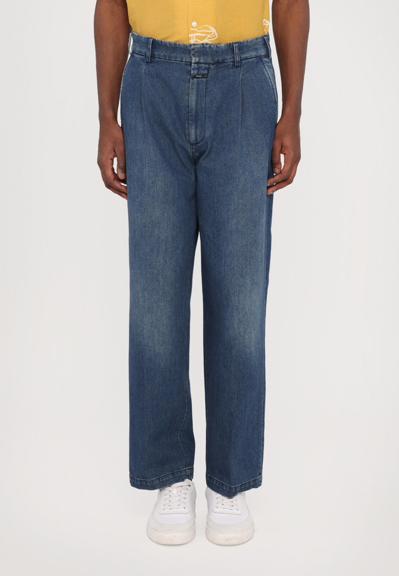 Blå denimbukser med en afslappet pasform, der har folder, en knaplukning og et kontrastfarvet liv. Båret med hvide sneakers.