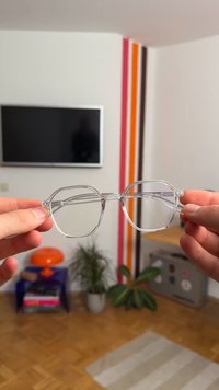 Selkeät, oktaalimuotoiset lasit, joita pidetään sormilla, ohuella kehykset ja huomaamattomat metallikorostukset temppeleissä. Taustalla minimalistinen sisustus.