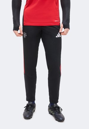 Personne portant un pantalon de survêtement Adidas Manchester United noir, des chaussures de football noires et un haut de sport rouge avec des manches noires.