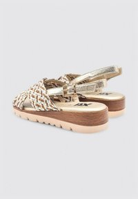 Sandalias trenzadas con correas doradas, que presentan un diseño entrelazado en tonos marrón y blanco. Suela de plataforma marrón con una superficie texturizada. Hebilla ajustable.