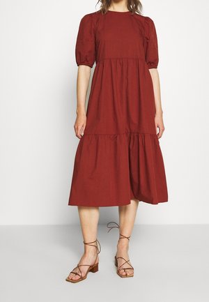 Femme portant une robe mi-longue rouge rouille avec des manches bouffantes et une jupe à volants, associée à des sandales marron à talons blocs et à brides.