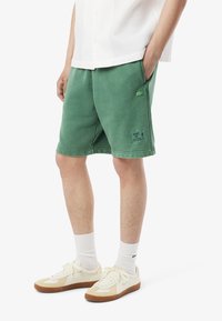 Shorts verts en mélange de coton avec une coupe décontractée, poches avant et un patch logo à l'ourlet. Associés à des baskets crème et des chaussettes blanches.