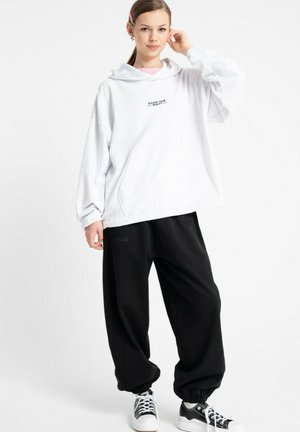 Biała, oversize'owa bluza z kapturem z napisem „Podnieś swoje piękno”, w połączeniu z czarnymi, luźnymi spodniami oraz czarno-białymi sneakersami. Prosty design, casualowy krój.