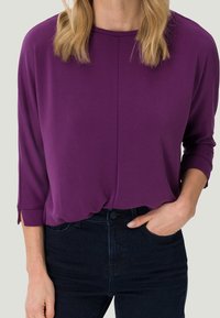 Frau in einer lila Langarmbluse, die in eine dunkelblaue Jeans gesteckt ist, mit einer Hand in ihrer Tasche vor einem neutralen Hintergrund.