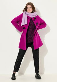 Pink uldblanding frakke med toggle-lukning, parret med et lilla ribbet tørklæde og sorte jeans, der har hvid side-stikning og kraftige sorte støvler.