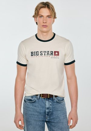 Młody mężczyzna w kremowym T-shircie z czarnym obszyciem i napisem "BIG STAR", ubrany w niebieskie dżinsy i brązowy pasek, stoi na białym tle.