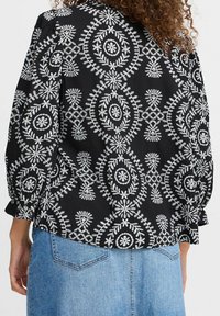 Blouse noire à motifs avec broderie florale blanche ; présente une coupe décontractée, des manches 3/4 et un ourlet arrondi. Associée à une jupe en denim.