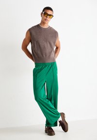 Pull sans manches marron, pantalon vert avec des accents de couture sur les côtés et chaussures marron. Tissu lisse avec une coupe décontractée et un design athlétique.