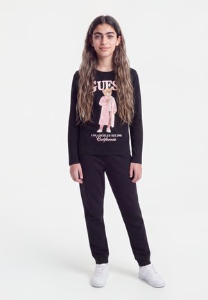 Jeune fille aux longs cheveux debout portant un sweat-shirt noir avec un motif d’ours et un texte, un pantalon noir et des baskets blanches sur fond uni.