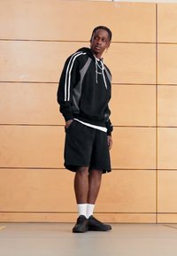 Camisola de capuz preta com detalhes brancos e painéis cinzentos, combinada com calções pretos e sapatilhas pretas. O cenário inclui uma parede de madeira.